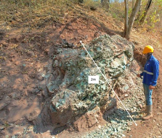 The picture shows a person in blue work clothes and a yellow helmet standing next to a large, exposed rock outcrop on which a measuring stick with a 2-meter mark has been placed.