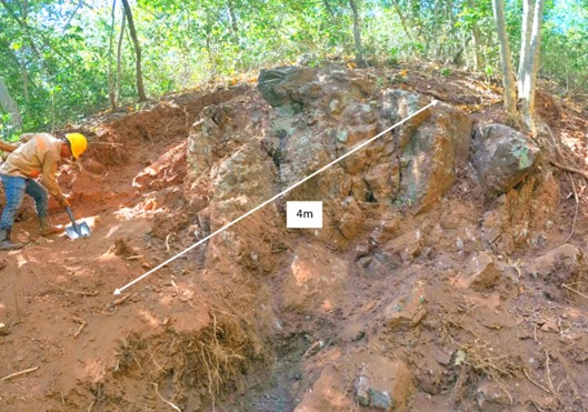 Ein Arbeiter in Schutzausrüstung untersucht einen großen, freigelegten Erzblock in einer Grube im Wald, neben ihm liegt ein Werkzeug. Ein Maßstab zeigt die 4 Meter breite Struktur.