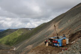 Goldbohrgerät an steilem Berghang mit Geröll, verhangener Himmel, im Hintergrund gründe Berglandschaft
